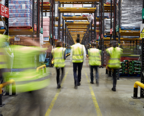 Staff In Reflective Vests Walking In A Warehouse, Back View